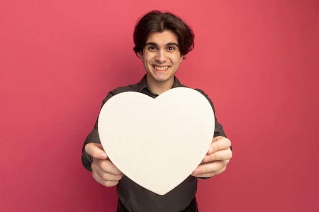 Chico guapo joven sonriente con camiseta negra sosteniendo una caja en forma de corazón en la parte delantera aislada en la pared rosa