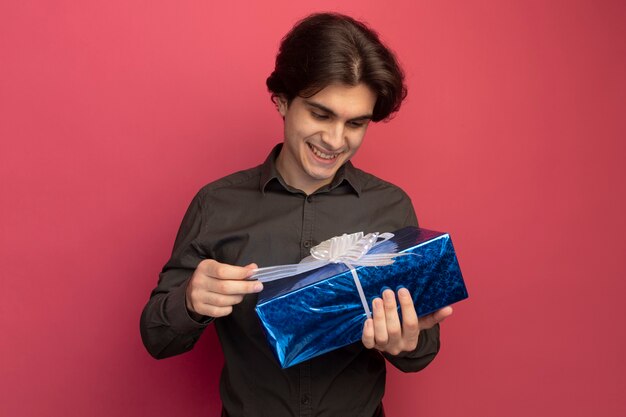 Chico guapo joven sonriente con camiseta negra sosteniendo y mirando la caja de regalo aislada en la pared rosa