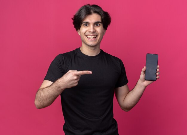 Chico guapo joven sonriente con camiseta negra sosteniendo y puntos en el teléfono aislado en la pared rosa