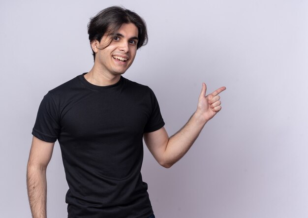 Chico guapo joven sonriente con puntos de camiseta negra en el lado aislado en la pared blanca con espacio de copia