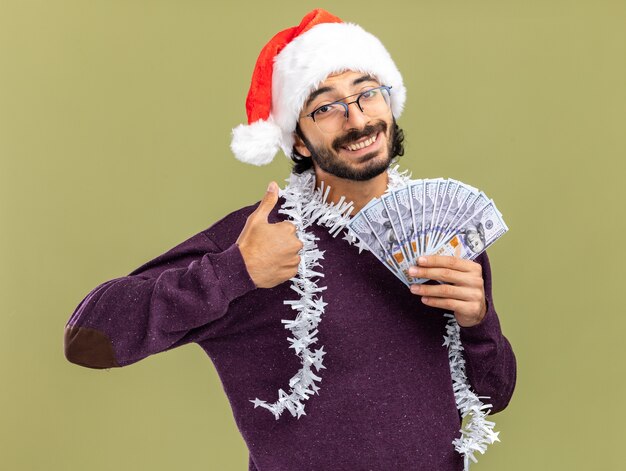 Chico guapo joven sonriente con sombrero de navidad con guirnalda en el cuello sosteniendo dinero en efectivo mostrando el pulgar hacia arriba aislado sobre fondo verde oliva