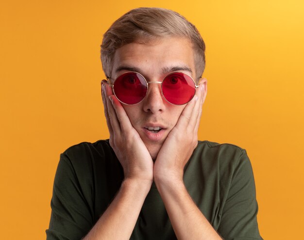 Chico guapo joven sorprendido con camisa verde con gafas poniendo las manos en las mejillas aisladas en la pared amarilla