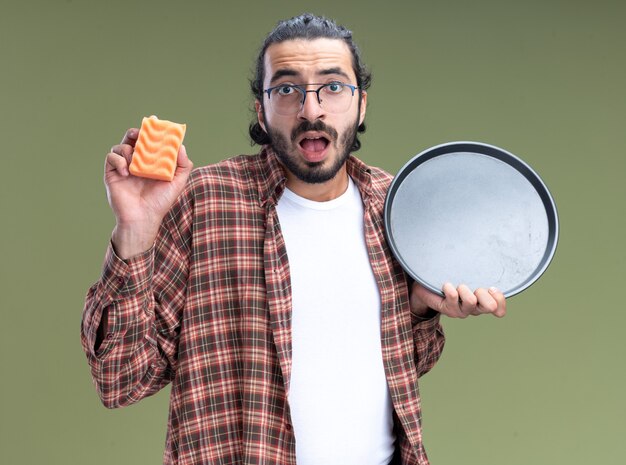 Foto gratuita chico guapo joven sorprendido de la limpieza que lleva la camiseta que sostiene la bandeja con la esponja aislada en la pared verde oliva