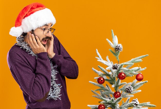 Chico guapo joven sorprendido de pie cerca del árbol de Navidad con sombrero de Navidad con guirnalda en el cuello poniendo la mano en las mejillas aisladas sobre fondo naranja
