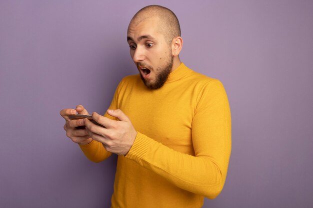 Chico guapo joven sorprendido sosteniendo y mirando el teléfono aislado en púrpura