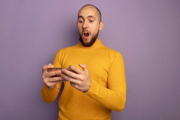 Chico guapo joven sorprendido sosteniendo y mirando el teléfono aislado en púrpura