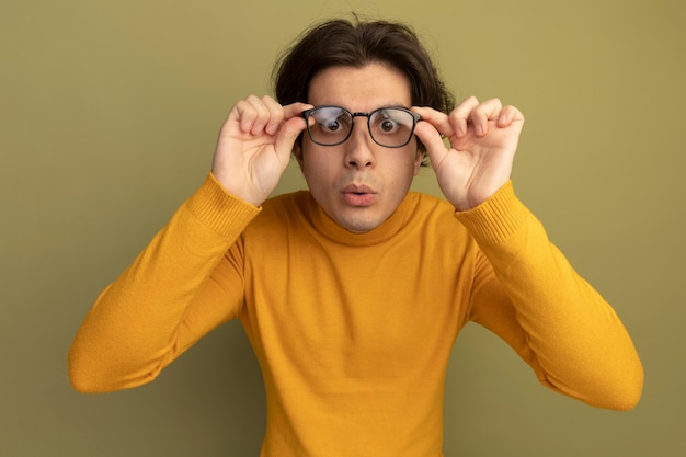 Chico guapo joven sorprendido con suéter de cuello alto amarillo y gafas aisladas en la pared verde oliva