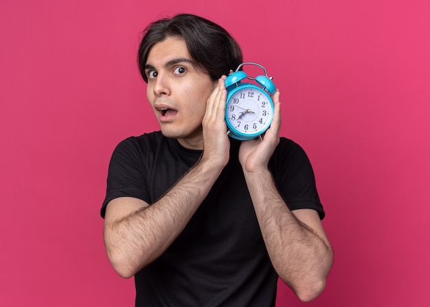 Chico guapo joven sospechoso con camiseta negra con reloj despertador alrededor de la oreja aislado en la pared rosa