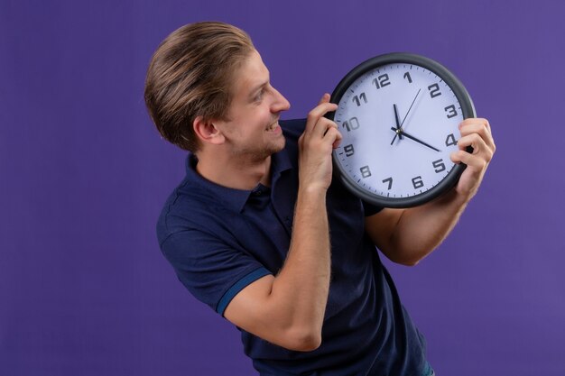 Chico guapo joven sosteniendo el reloj en las manos mirándolo con cara feliz salido y alegre de pie sobre fondo púrpura