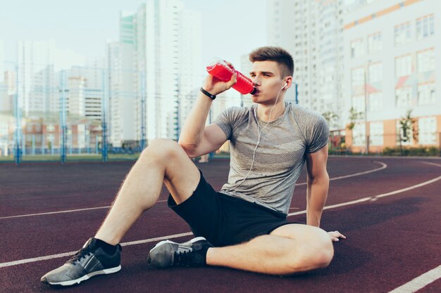 Chico guapo joven tiene un descanso del entrenamiento en el estadio por la mañana. Viste ropa deportiva, escucha música a través de auriculares, bebe bebida roja de la botella.