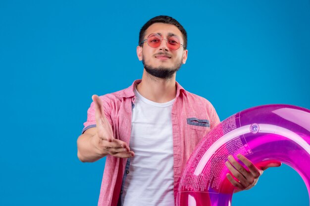 Chico guapo joven viajero con gafas de sol con anillo inflable mirando a la cámara con una sonrisa ofreciendo mano haciendo gesto de saludo sobre fondo azul