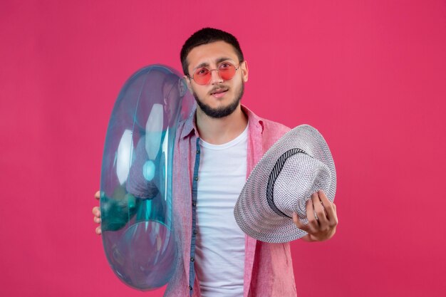 Chico guapo joven viajero con gafas de sol con anillo inflable y sombrero de verano mirando confundido de pie sobre fondo rosa