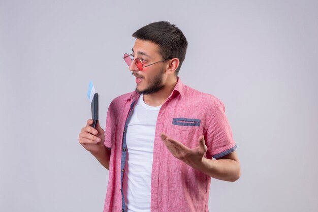 Chico guapo joven viajero con gafas de sol con boletos aéreos mirando a un lado discutiendo con alguien de pie sobre fondo blanco