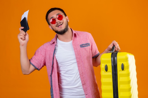 Chico guapo joven viajero con gafas de sol con maleta y boletos aéreos mirando seguro de sí mismo satisfecho sonriendo alegremente listo para viajar de pie sobre fondo naranja