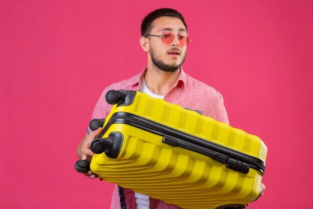 Chico guapo joven viajero con gafas de sol con maleta mirando a un lado con expresión de confianza en la cara de pie sobre fondo rosa