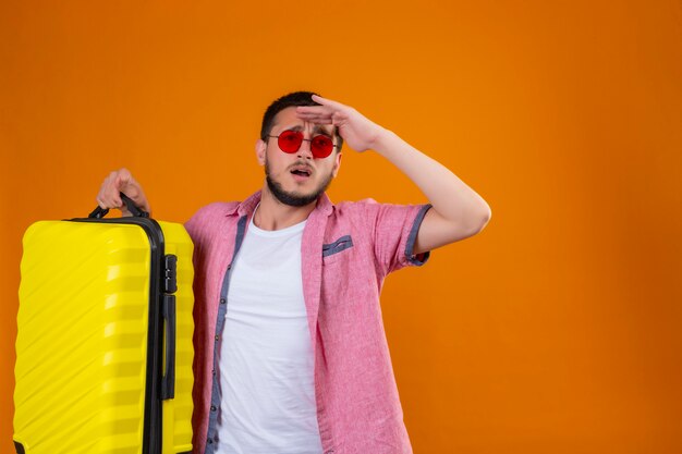 Chico guapo joven viajero con gafas de sol con maleta mirando lejos con la mano para mirar algo con expresión confusa de pie sobre fondo naranja