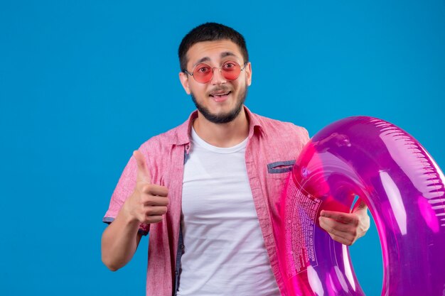 Chico guapo joven viajero con gafas de sol sosteniendo un anillo inflable mirando alegre y feliz sonriendo alegremente mostrando los pulgares para arriba sobre fondo azul