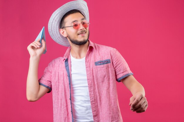 Chico guapo joven viajero en sombrero de verano con gafas de sol mirando seguro lanzando avión de papel de pie sobre fondo rosa