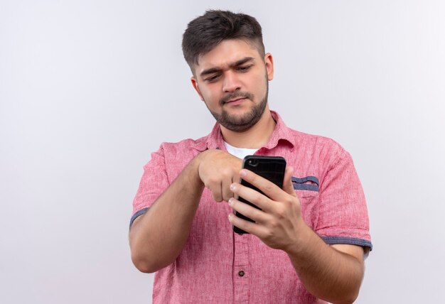 Chico guapo joven vistiendo polo rosa apuntando en el teléfono de pie sobre la pared blanca