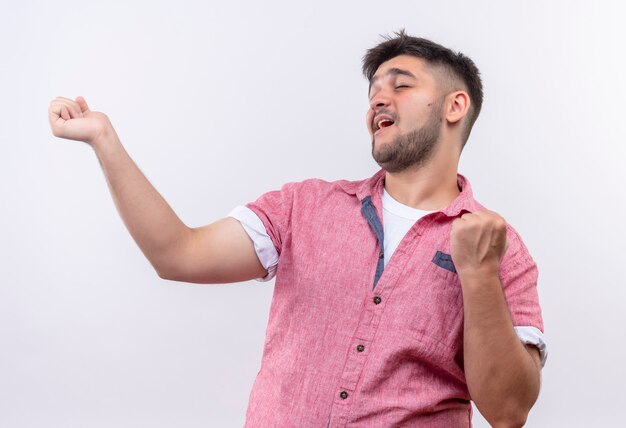 Chico guapo joven vistiendo polo rosa bailando sus puños de pie sobre la pared blanca