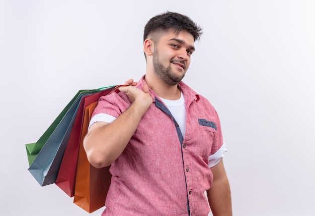 Foto gratuita chico guapo joven vistiendo polo rosa contento mirando sosteniendo bolsas en la espalda de pie sobre la pared blanca