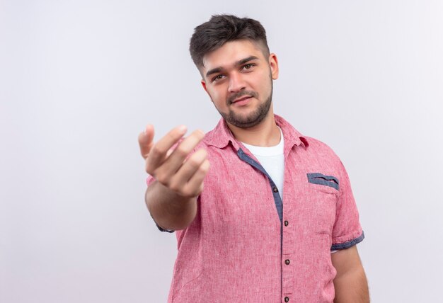 Chico guapo joven vistiendo polo rosa cordialmente llamándolo parado sobre la pared blanca