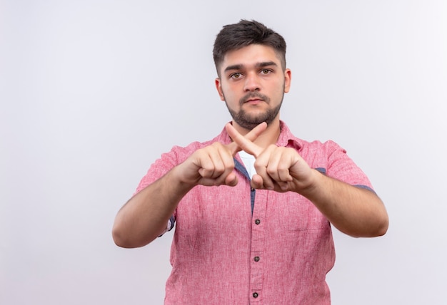 Chico guapo joven vistiendo polo rosa cruzando el dedo seriamente parado sobre la pared blanca