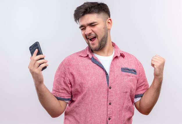 Chico guapo joven vistiendo polo rosa felizmente cerrando los ojos sosteniendo el teléfono parado sobre la pared blanca