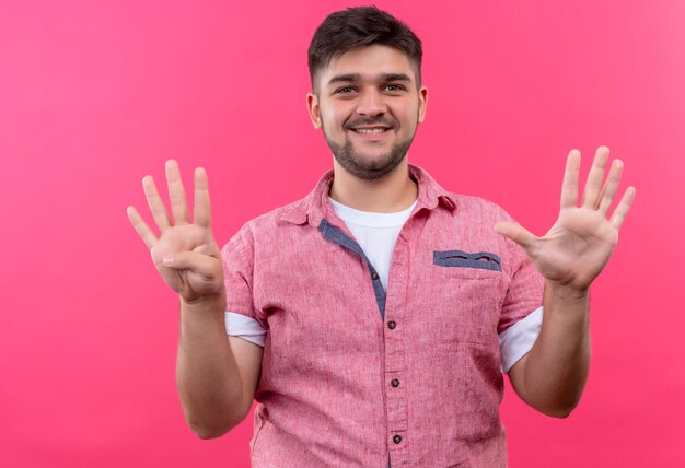 Chico guapo joven vistiendo polo rosa felizmente mostrando nueve signos con los dedos de pie sobre la pared rosa