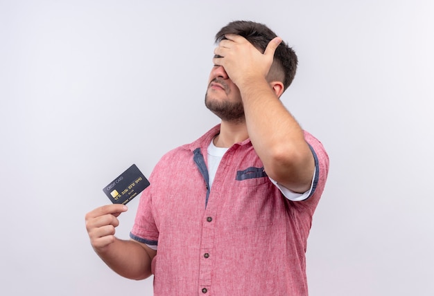 Foto gratuita chico guapo joven vistiendo polo rosa haciendo facepalm molesto sosteniendo la tarjeta de crédito de pie sobre la pared blanca