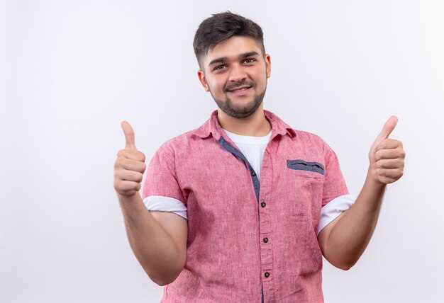 Chico guapo joven vistiendo polo rosa haciendo felices pulgares para arriba de pie sobre la pared blanca