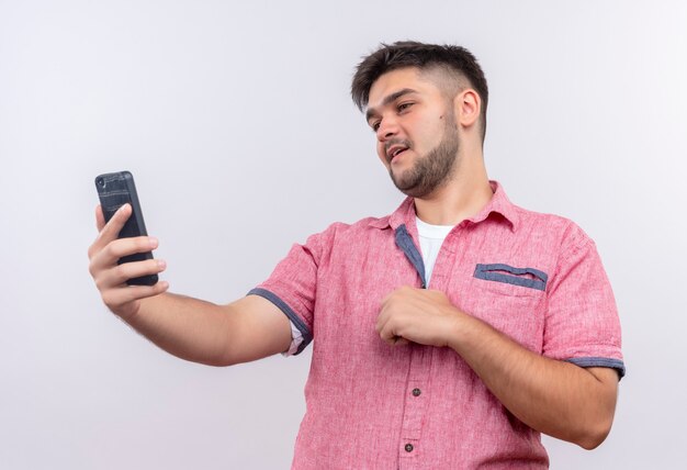 Chico guapo joven vistiendo polo rosa haciendo selfie complacido de pie sobre la pared blanca