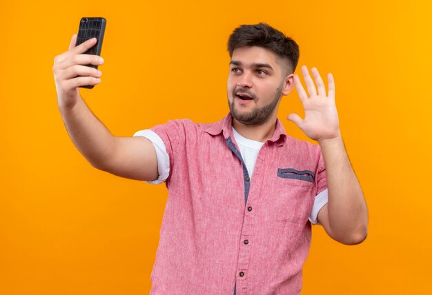 Chico guapo joven vistiendo polo rosa haciendo selfie diciendo hola de pie sobre la pared naranja