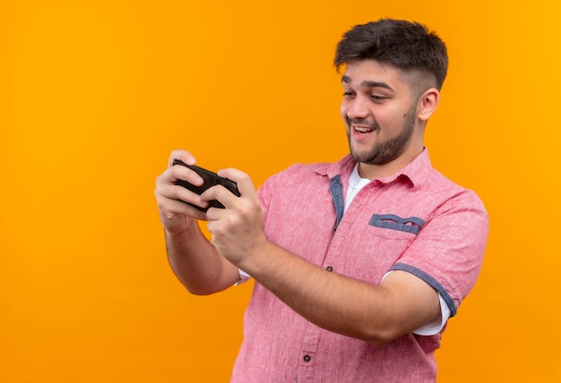 Foto gratuita chico guapo joven vistiendo polo rosa jugando felizmente en el teléfono de pie sobre la pared naranja