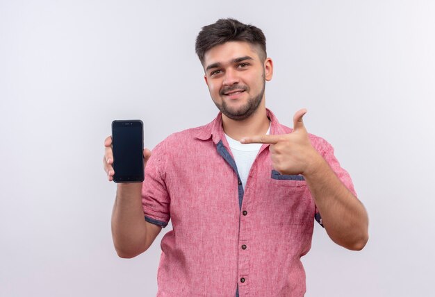 Chico guapo joven vistiendo polo rosa mirando felizmente apuntando al teléfono con el dedo índice de pie sobre la pared blanca