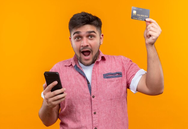 Chico guapo joven vistiendo polo rosa mirando felizmente sosteniendo el teléfono levantando la tarjeta de crédito de pie sobre la pared naranja