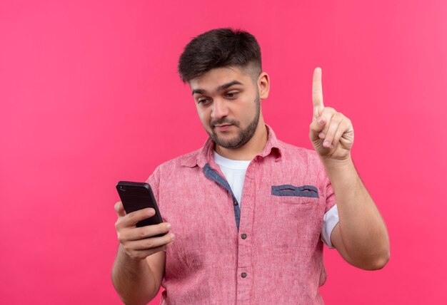 Chico guapo joven vistiendo polo rosa mirando teléfono sorprendido apuntando con el dedo índice de pie sobre la pared rosa