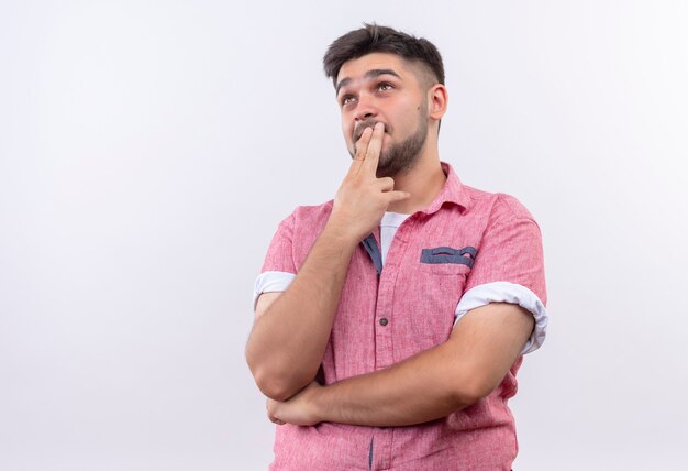 Chico guapo joven vistiendo polo rosa pensativamente mirando hacia arriba tocando la boca con los dedos de pie sobre la pared blanca