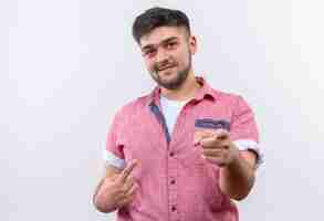 Foto gratuita chico guapo joven vistiendo polo rosa sonriendo señalando con los dedos índices de pie sobre la pared blanca