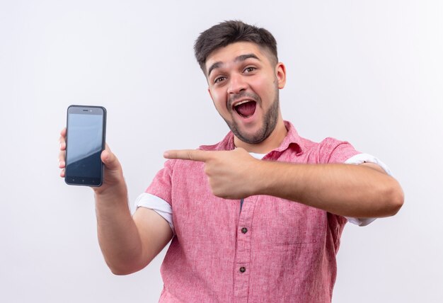 Chico guapo joven vistiendo polo rosa sorprendido apuntando al teléfono con el dedo índice izquierdo parado sobre la pared blanca