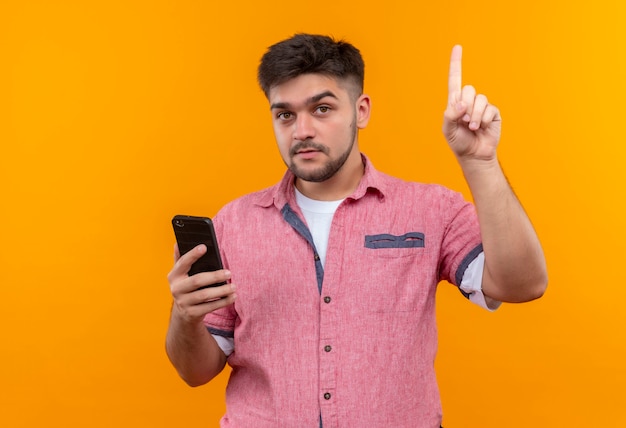 Foto gratuita chico guapo joven vistiendo polo rosa sosteniendo el teléfono concentrado apuntando hacia arriba con el dedo índice de pie sobre la pared naranja