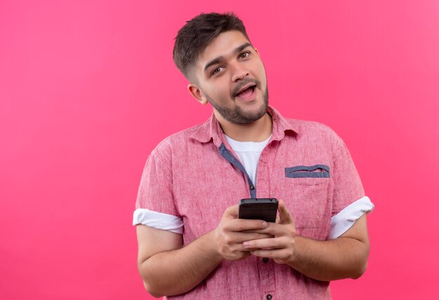 Chico guapo joven vistiendo polo rosa sosteniendo teléfono juguetonamente mirando parado sobre pared rosa