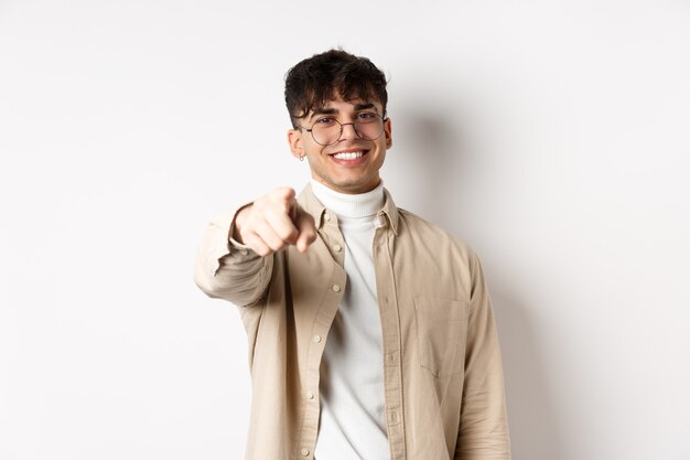 Chico guapo moderno con gafas apuntando a la cámara, sonriendo y eligiéndote, reclutando o invitando al evento, de pie sobre fondo blanco.