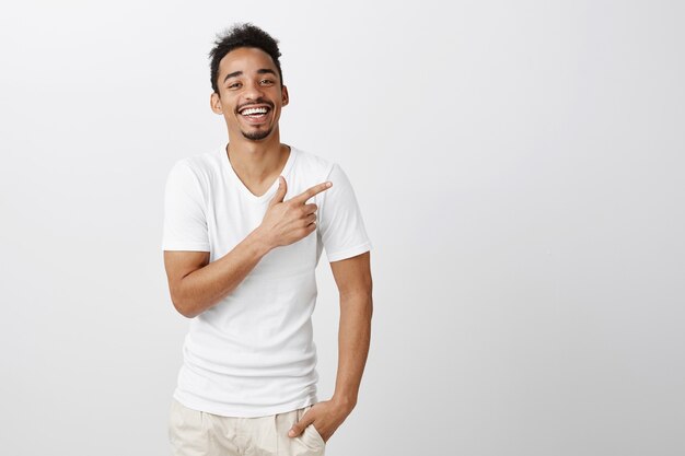 Chico guapo de piel oscura complacido en camiseta blanca apuntando hacia la esquina superior derecha, haciendo una elección