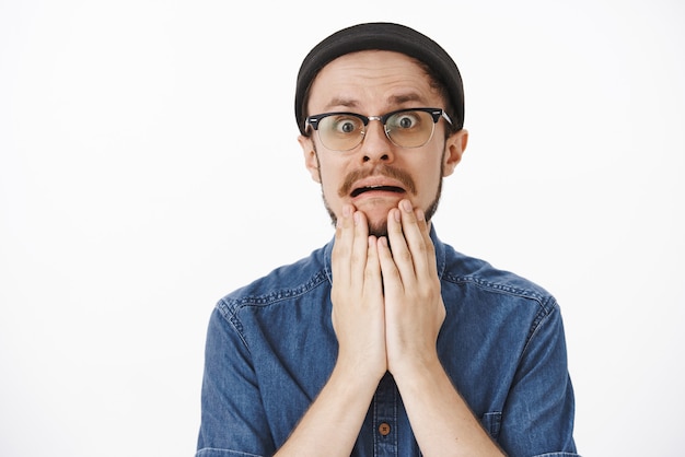 Chico guapo con problemas desesperados con barba en gorro negro y gafas sosteniendo las palmas en la barbilla frunciendo el ceño y haciendo muecas de pánico mientras se encuentra en una situación terrible mirando asustado sobre la pared gris