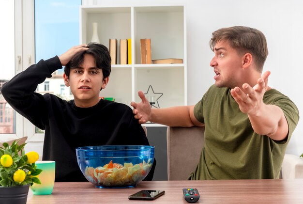 El chico guapo rubio joven molesto apunta a la cámara con la mano sentada en la mesa y mirando al chico guapo moreno joven decepcionado poniendo la mano en la cabeza mirando a la cámara dentro de la sala de estar