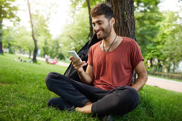 Chico guapo sentado en el parque, apoyado en el árbol y usando el teléfono móvil, desplácese por la aplicación de redes sociales, charlando