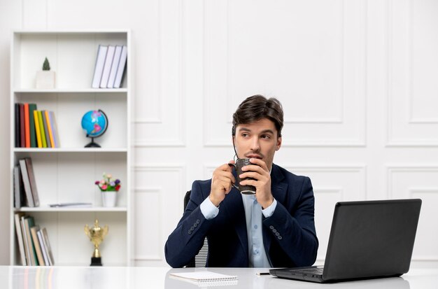 Chico guapo de servicio al cliente con auriculares y computadora portátil en traje bebiendo de una taza