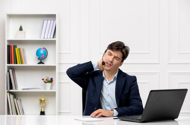 Chico guapo de servicio al cliente con auriculares y computadora portátil en traje cansado sosteniendo su cuello