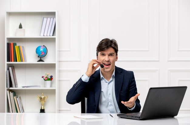 Chico guapo de servicio al cliente con auriculares y computadora portátil en traje molesto hablando con alguien